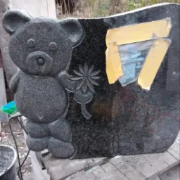 Granitowy nagrobek dziecięcy z płaskorzeźbą misia trzymającego kwiat, zabezpieczony taśmą z fragmentem zdjęcia, ustawiony na drewnianej palecie.