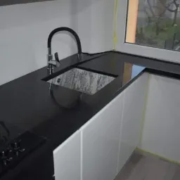 Czarny, błyszczący blat kuchenny z wbudowanym zlewem o marmurkowym wzorze i czarną baterią, obok płyta grzewcza; widoczne białe szafki i okno z czarnym parapetem.
