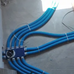 Niebieskie rury karbowane prowadzone od rozdzielacza do skrzynki podłączeniowej, zabezpieczone opaskami zaciskowymi, widoczne elementy instalacji w surowym stanie.