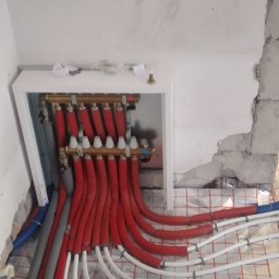 KANMAKS - Rozdzielacz ogrzewania podłogowego z czerwonymi rurami w izolacji, widoczne nieotynkowane fragmenty ściany oraz folia izolacyjna z nadrukowaną siatką.