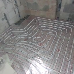 KANMAKS - Białe rury ogrzewania podłogowego rozłożone na folii z czerwoną siatką w surowym wnętrzu z ceglanymi ścianami w trakcie montażu.