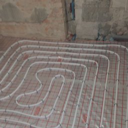 KANMAKS - Białe rury ogrzewania podłogowego rozłożone na folii z czerwoną siatką, widok na surową ścianę z fragmentami tynku.