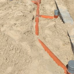 KANMAKS - Pomarańczowe rury kanalizacyjne ułożone w wykopie zasypanym piaskiem, widoczne studzienki kontrolne.