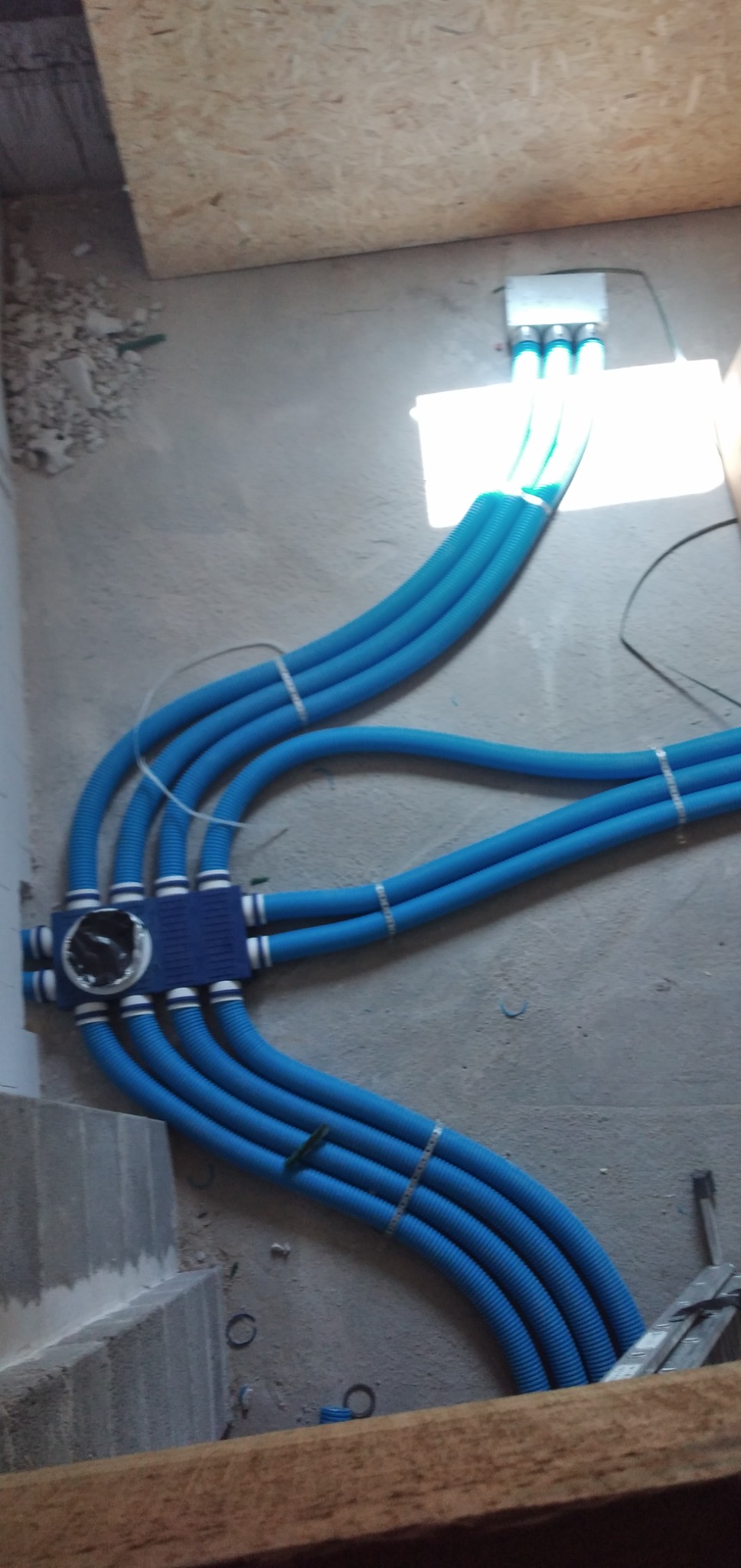 Niebieskie rury karbowane prowadzone od rozdzielacza do skrzynki podłączeniowej, zabezpieczone opaskami zaciskowymi, widoczne elementy instalacji w surowym stanie.