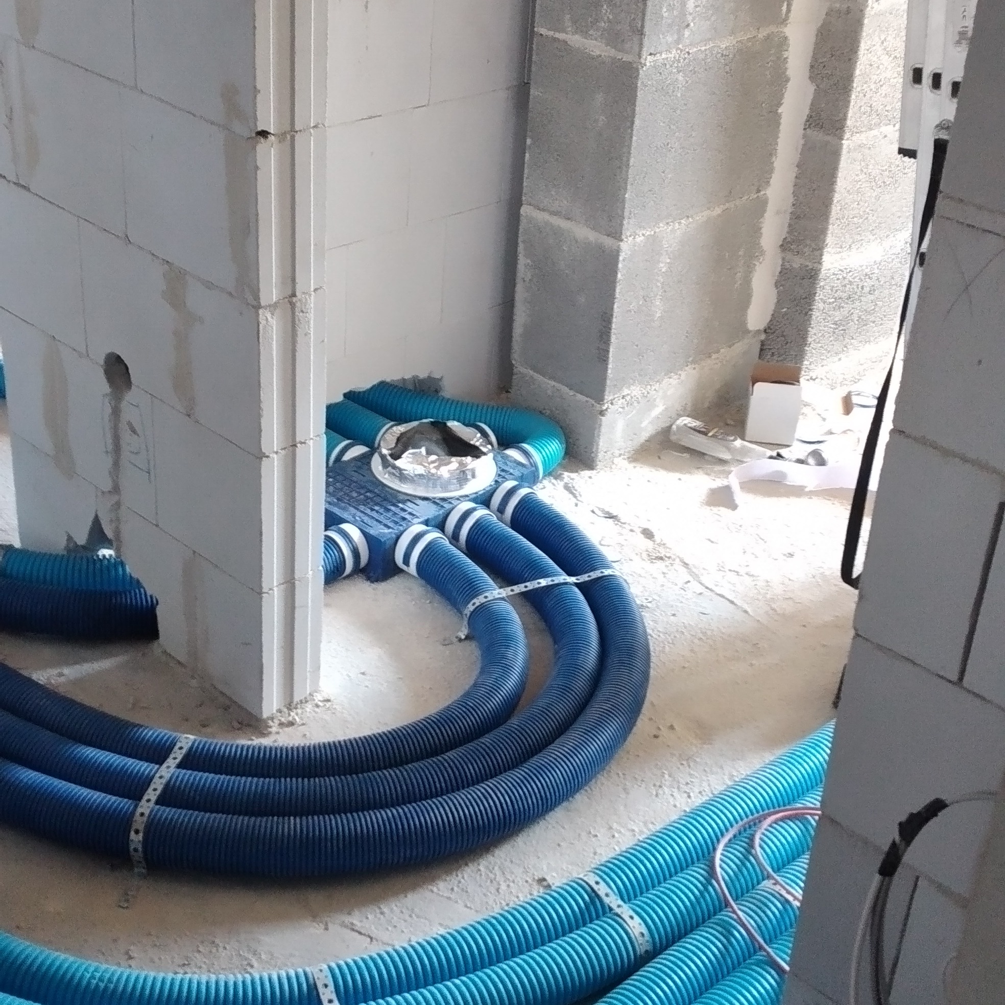 Niebieskie, karbowane rury wentylacyjne ułożone na podłodze w surowym wnętrzu, częściowo przymocowane metalową taśmą, z widocznymi bloczkami betonowymi w tle.