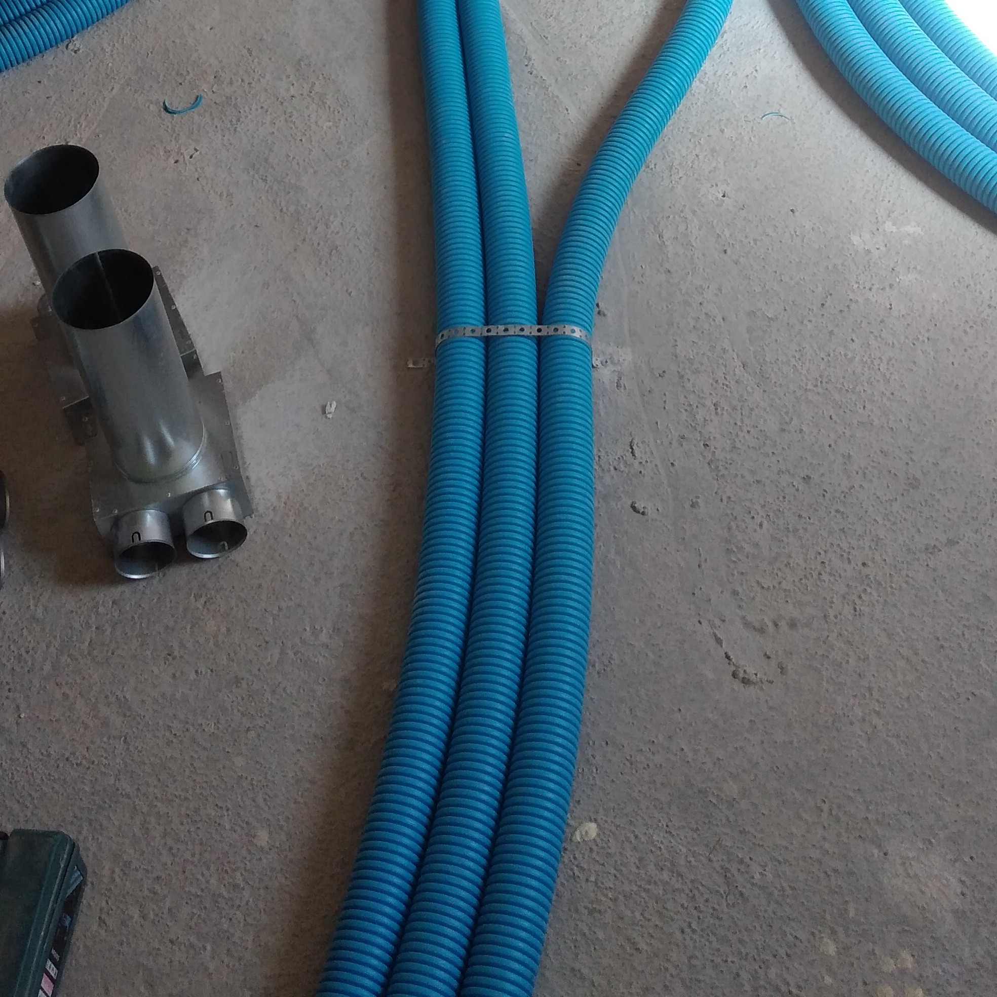 Niebieskie rury karbowane spięte metalową obejmą, obok metalowy rozdzielacz na surowym betonowym podłożu w trakcie montażu.