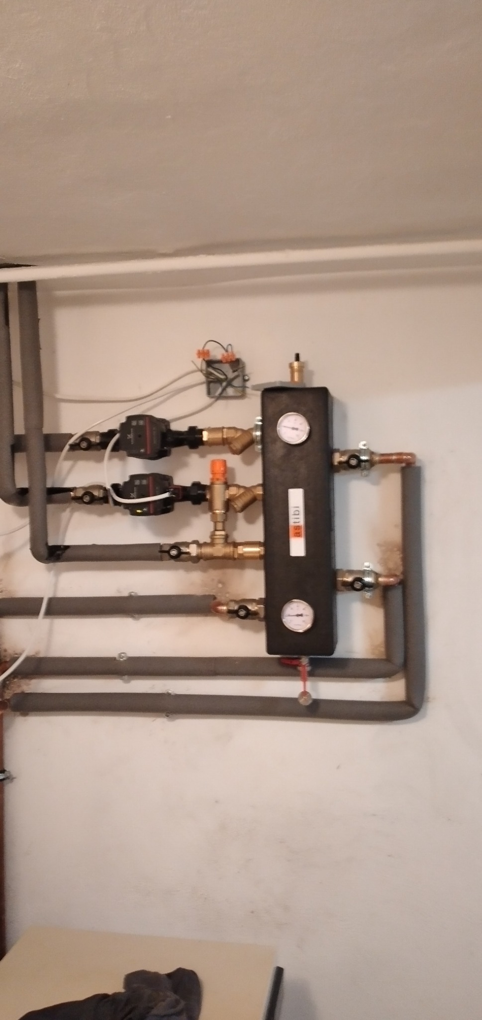 Rozdzielacz grzewczy Astibi z dwoma pompami obiegowymi na tle białej ściany, rury izolowane szarą pianką, manometry wskazują ciśnienie.