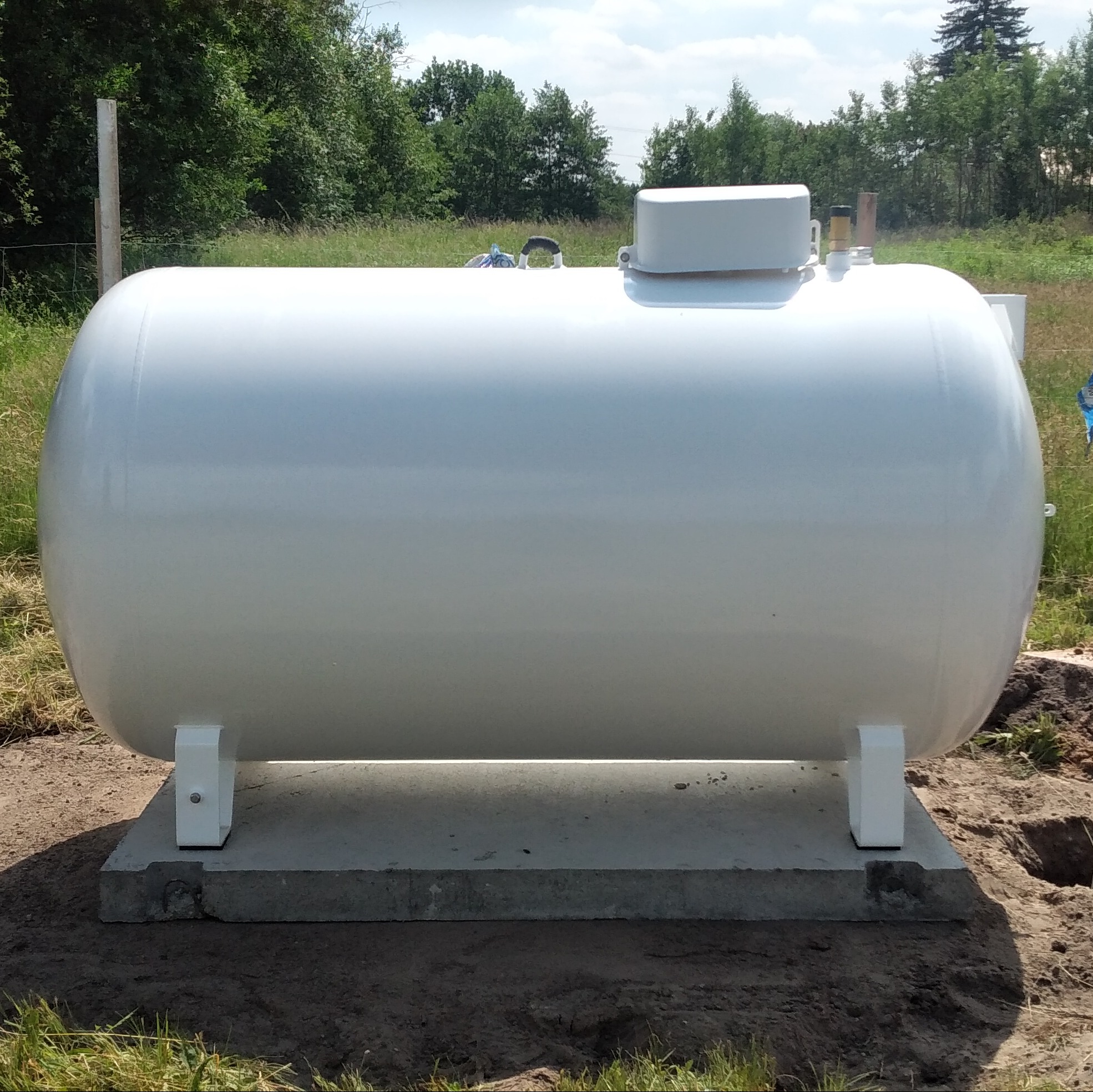 Biały, poziomy zbiornik gazu LPG na betonowej podstawie, widoczne zawory i przyłącza, tło stanowią drzewa i błękitne niebo.