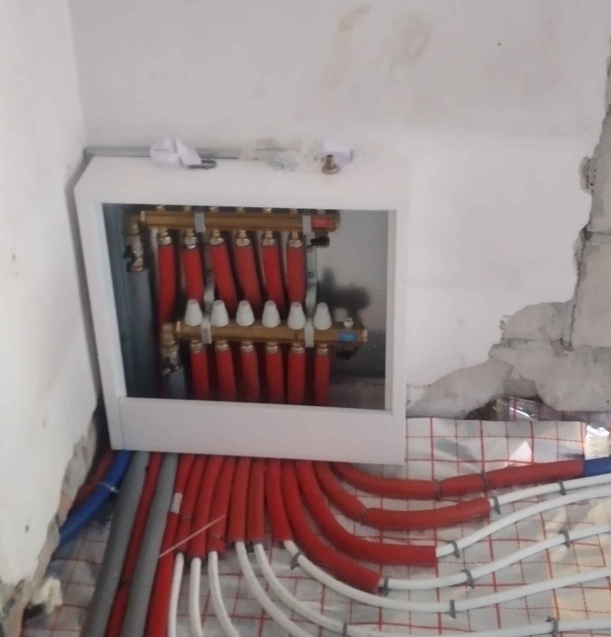 Rozdzielacz ogrzewania podłogowego z czerwonymi i białymi rurami w szafce, instalacja w trakcie montażu, widoczny styropian z rastrem pod rury.