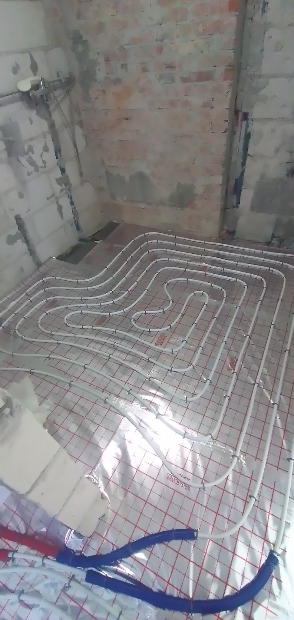 Białe rury ogrzewania podłogowego rozłożone na folii z czerwoną siatką w surowym wnętrzu z ceglanymi ścianami w trakcie montażu.