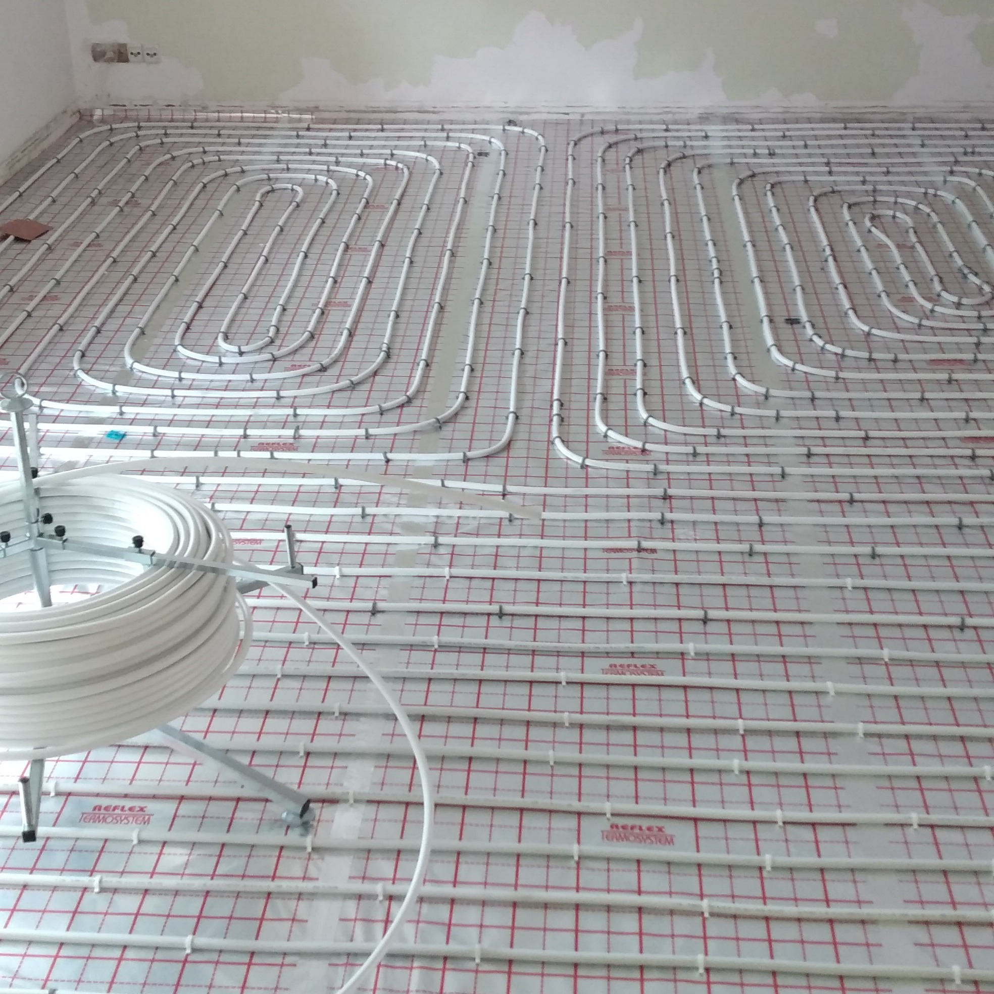 Rozłożona instalacja ogrzewania podłogowego na folii z siatką w pomieszczeniu w trakcie remontu, widoczny zwój rur na metalowym stojaku.