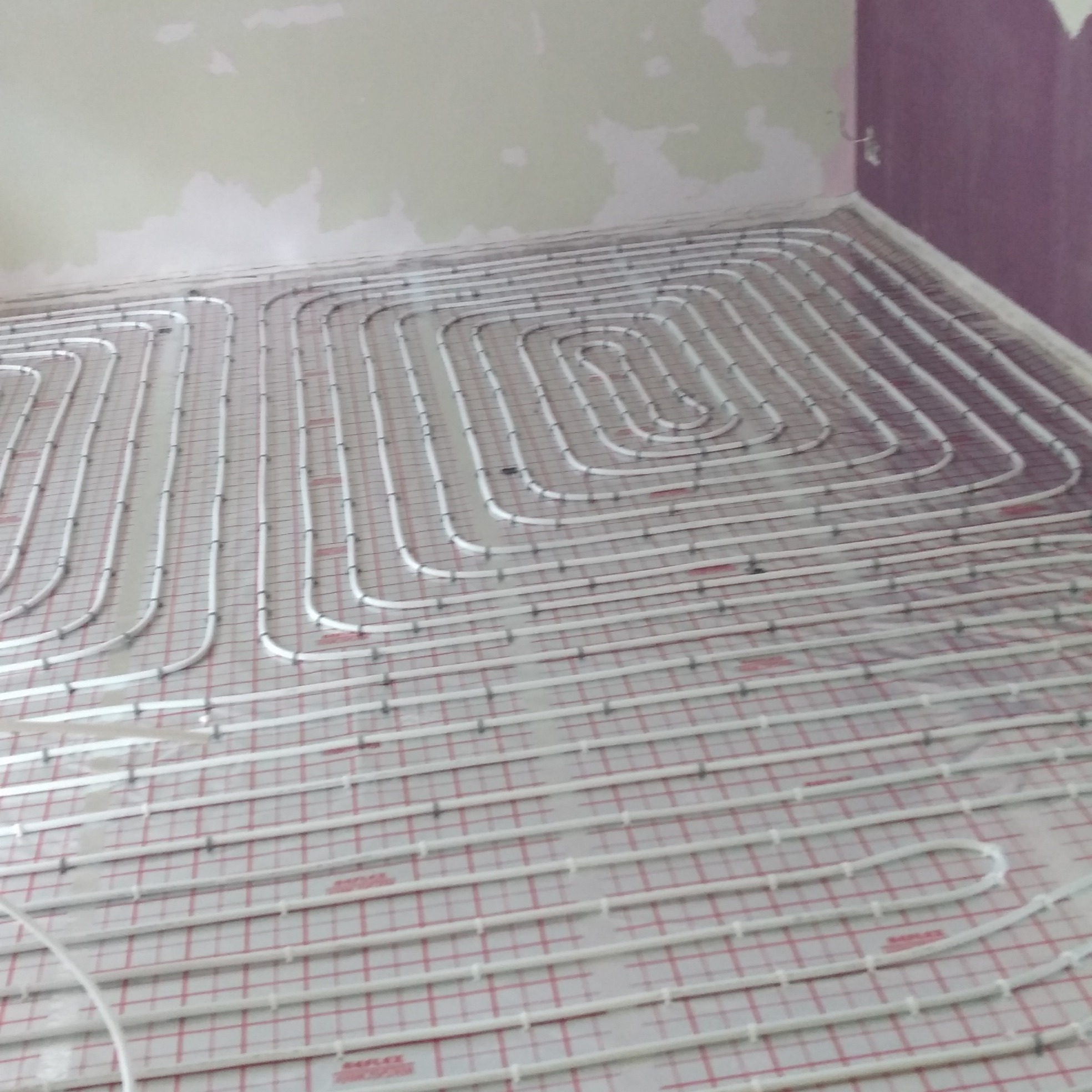 Białe rury ogrzewania podłogowego rozłożone na folii z czerwoną siatką, widok z góry na niedokończoną instalację w pomieszczeniu z surowymi ścianami.