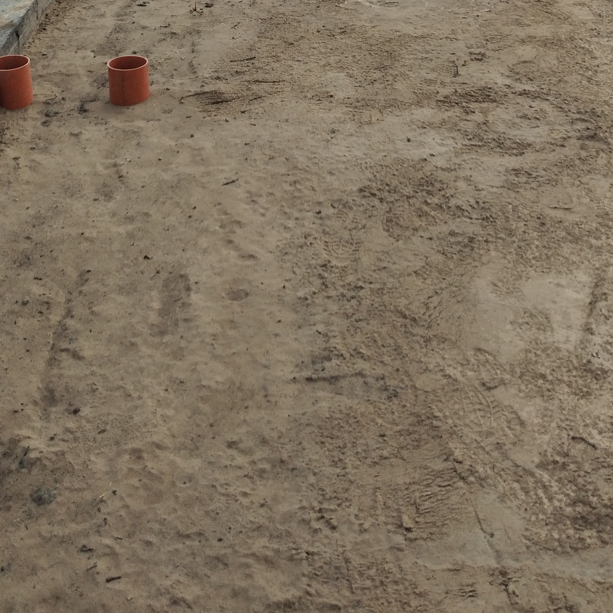 Dwa pomarańczowe rury kanalizacyjne leżące na piasku z widocznymi śladami stóp.