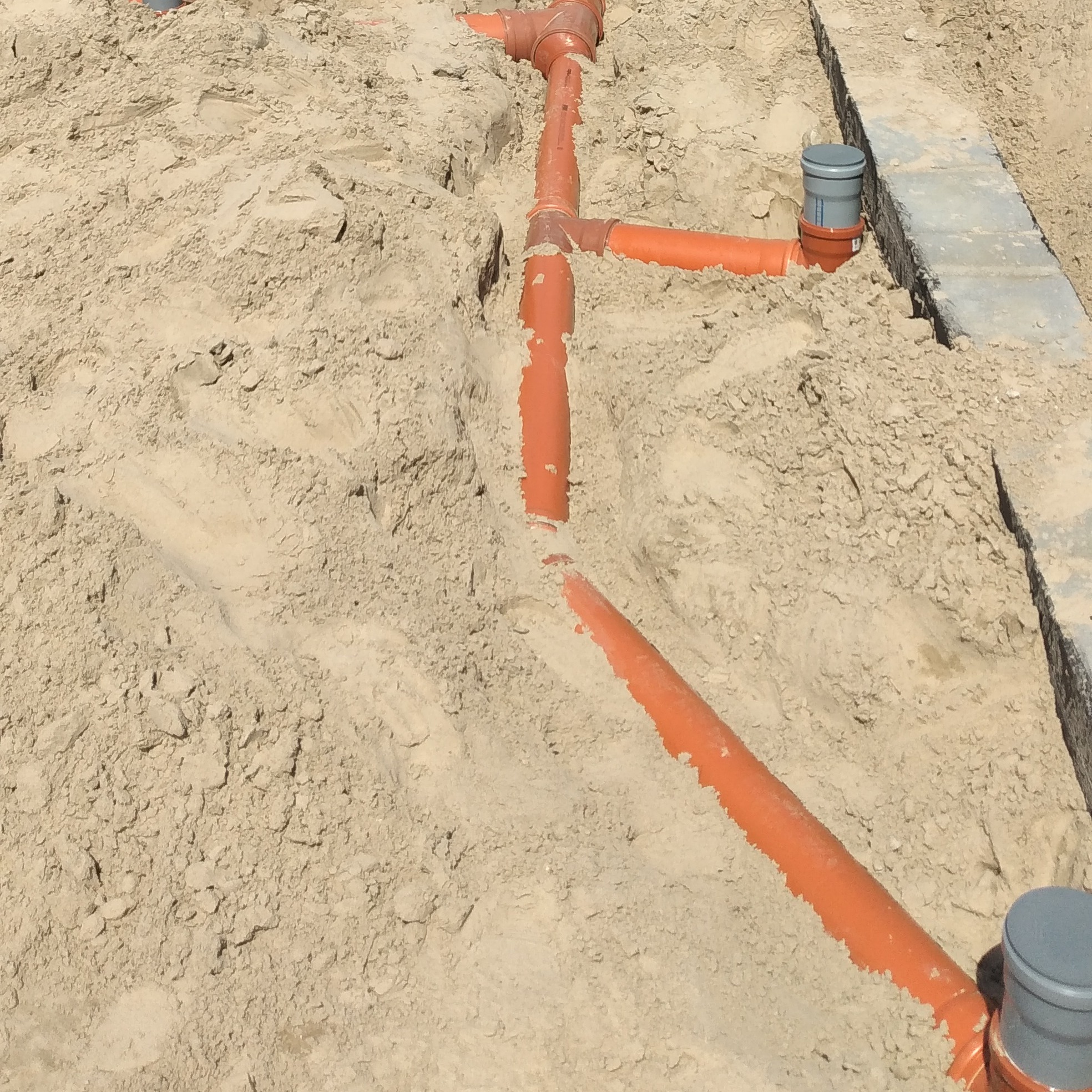 Pomarańczowe rury kanalizacyjne ułożone w wykopie zasypanym piaskiem, widoczne studzienki kontrolne.