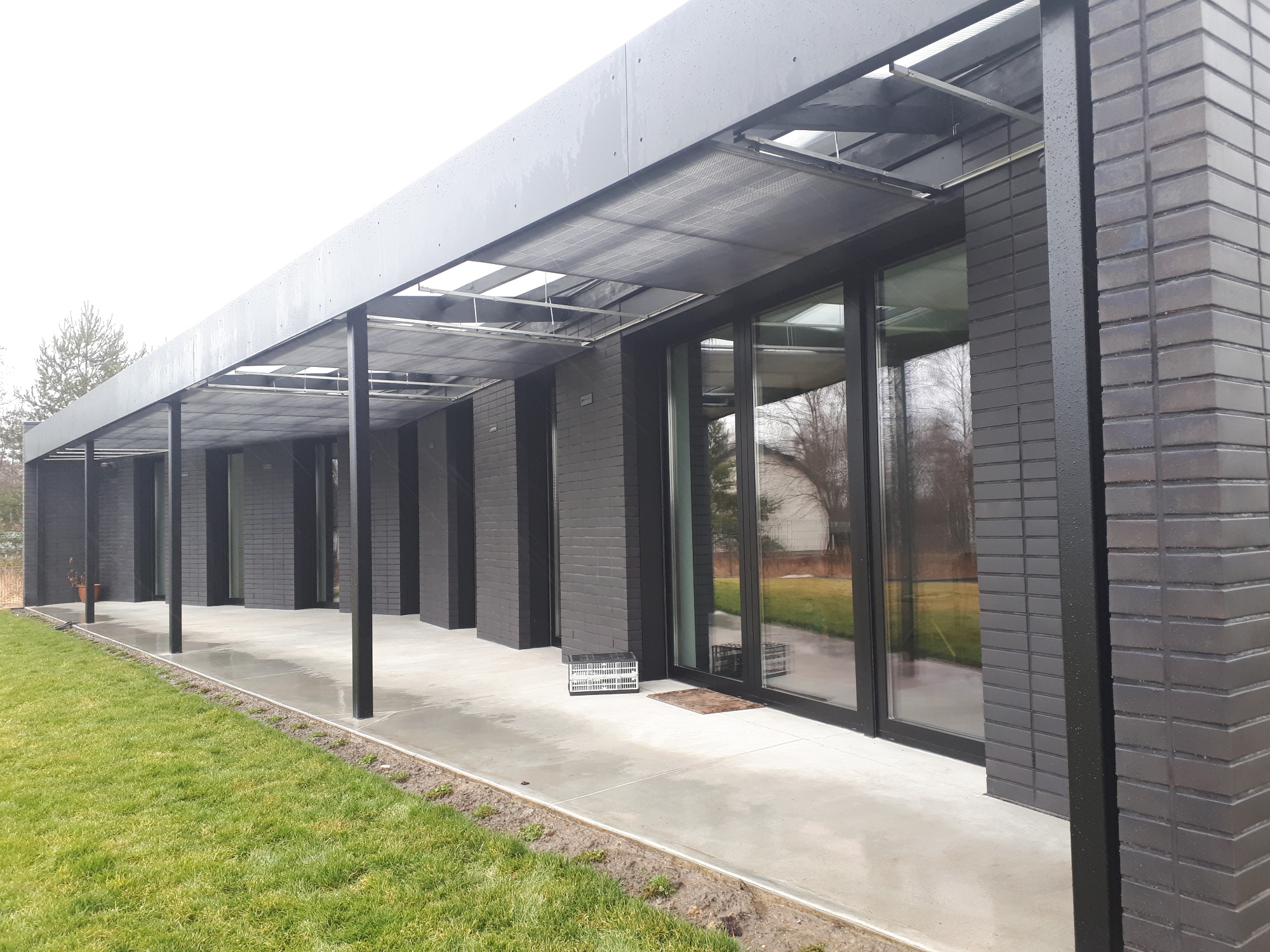 Nowoczesny dom z dużymi oknami w czarnych ramach i zadaszeniem w Tarnowskich Górach, widok z zewnątrz, zieleń trawnika.