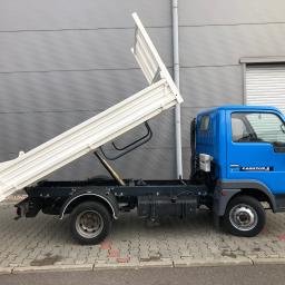 Gp-Partner - Niebieski samochód ciężarowy z otwartą, białą skrzynią ładunkową typu wywrotka, zaparkowany na kostce brukowej przed szarą ścianą budynku.