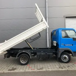 Niebieski samochód ciężarowy z otwartą, białą skrzynią ładunkową typu wywrotka, zaparkowany na kostce brukowej przed szarą ścianą budynku.