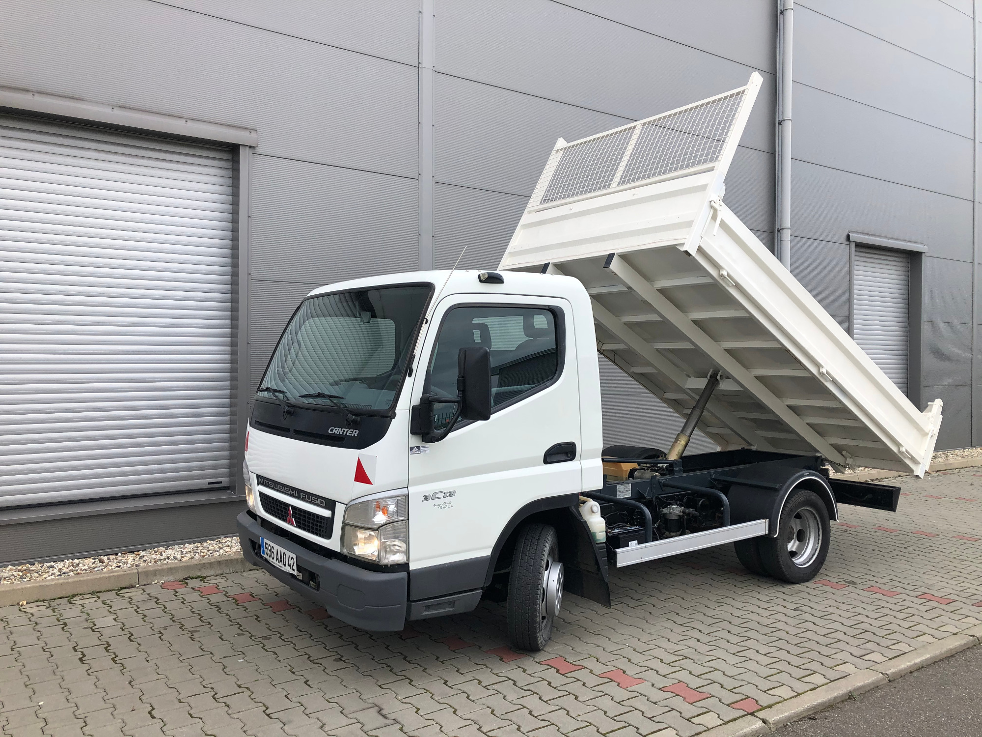 Biały samochód ciężarowy typu wywrotka Mitsubishi Fuso Canter z podniesioną skrzynią ładunkową, zaparkowany na kostce brukowej przed szarą halą przemysłową z bramą rolowaną.