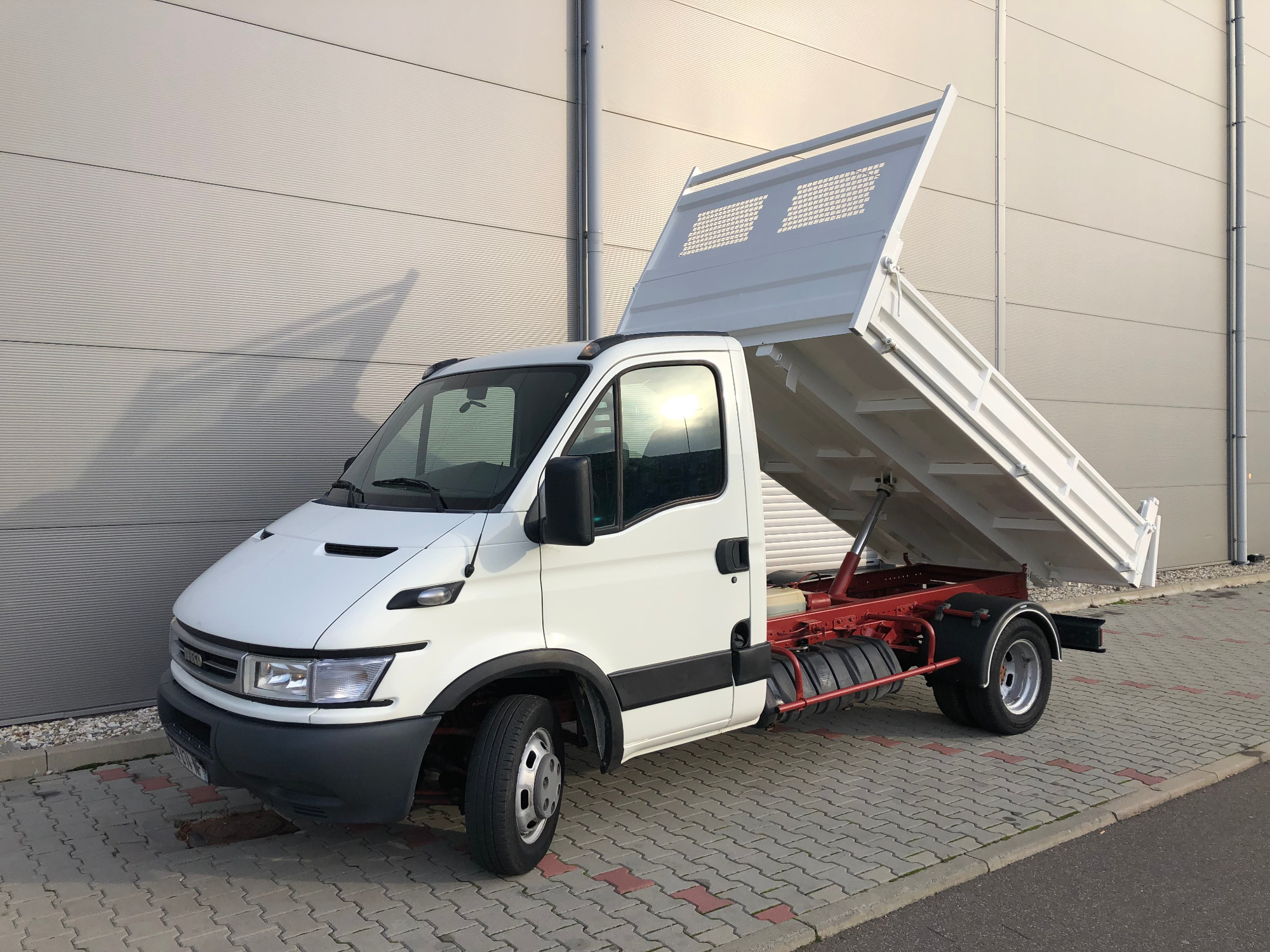 Biały samochód dostawczy typu wywrotka Iveco z podniesioną skrzynią ładunkową, zaparkowany na kostce brukowej przed budynkiem o szarej, panelowej elewacji.