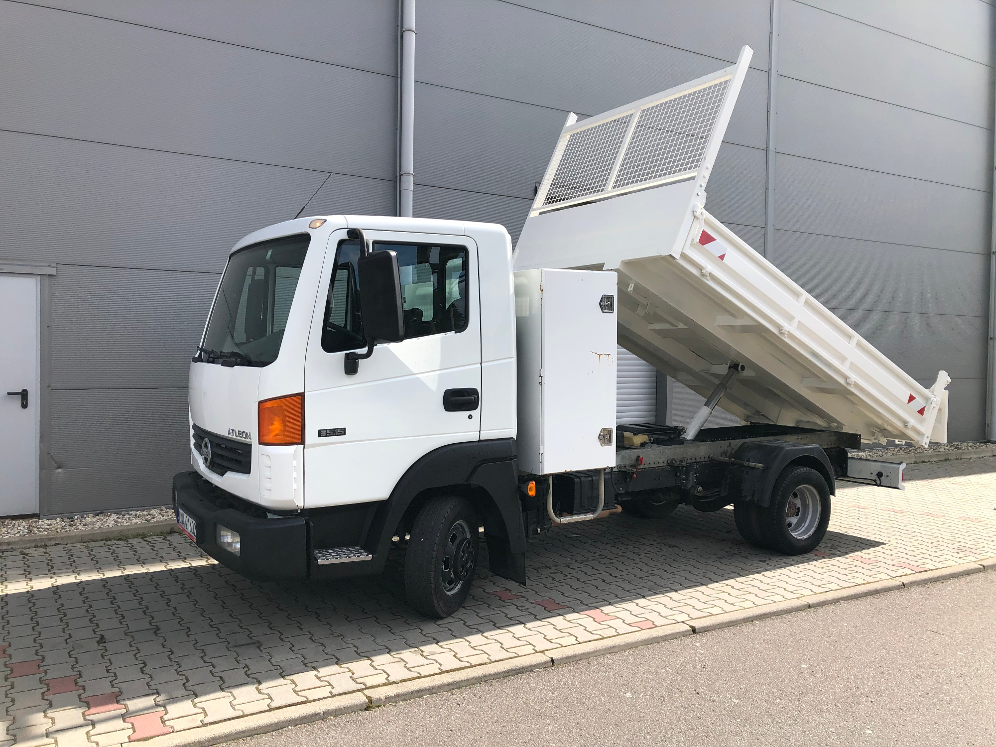 Biały samochód ciężarowy z otwartą skrzynią ładunkową, zaparkowany na kostce brukowej przed szarą ścianą budynku.