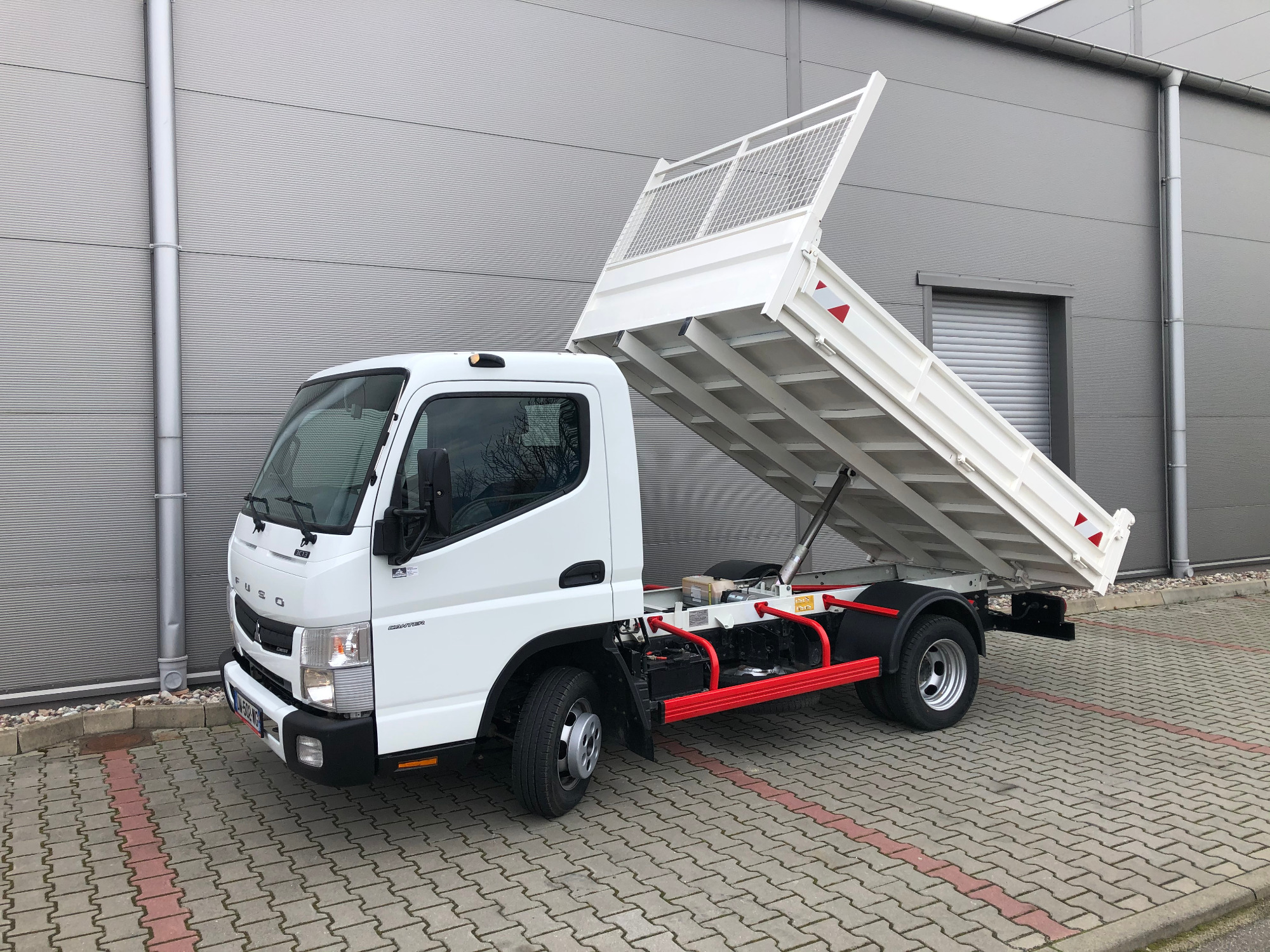 Biały wywrotny samochód ciężarowy Fuso Canter z podniesioną skrzynią ładunkową na tle szarej hali przemysłowej.