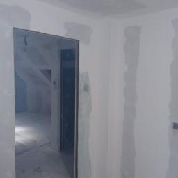 P.P.U.H Wojkoz - Wnętrze w trakcie remontu: ściany z płyt karton-gips, widoczne łączenia przed szpachlowaniem, surowe oświetlenie, widok na poddasze przez otwarte drzwi.