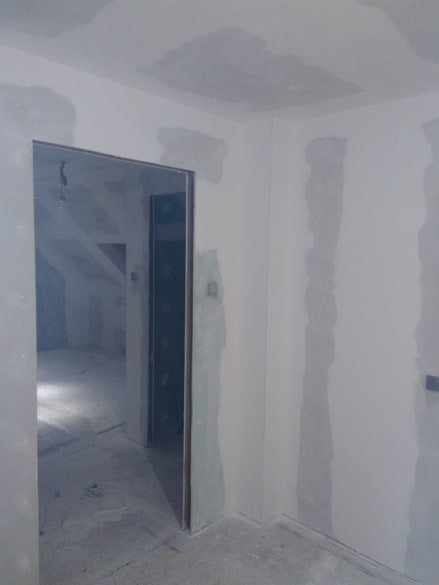 Wnętrze w trakcie remontu: ściany z płyt karton-gips, widoczne łączenia przed szpachlowaniem, surowe oświetlenie, widok na poddasze przez otwarte drzwi.