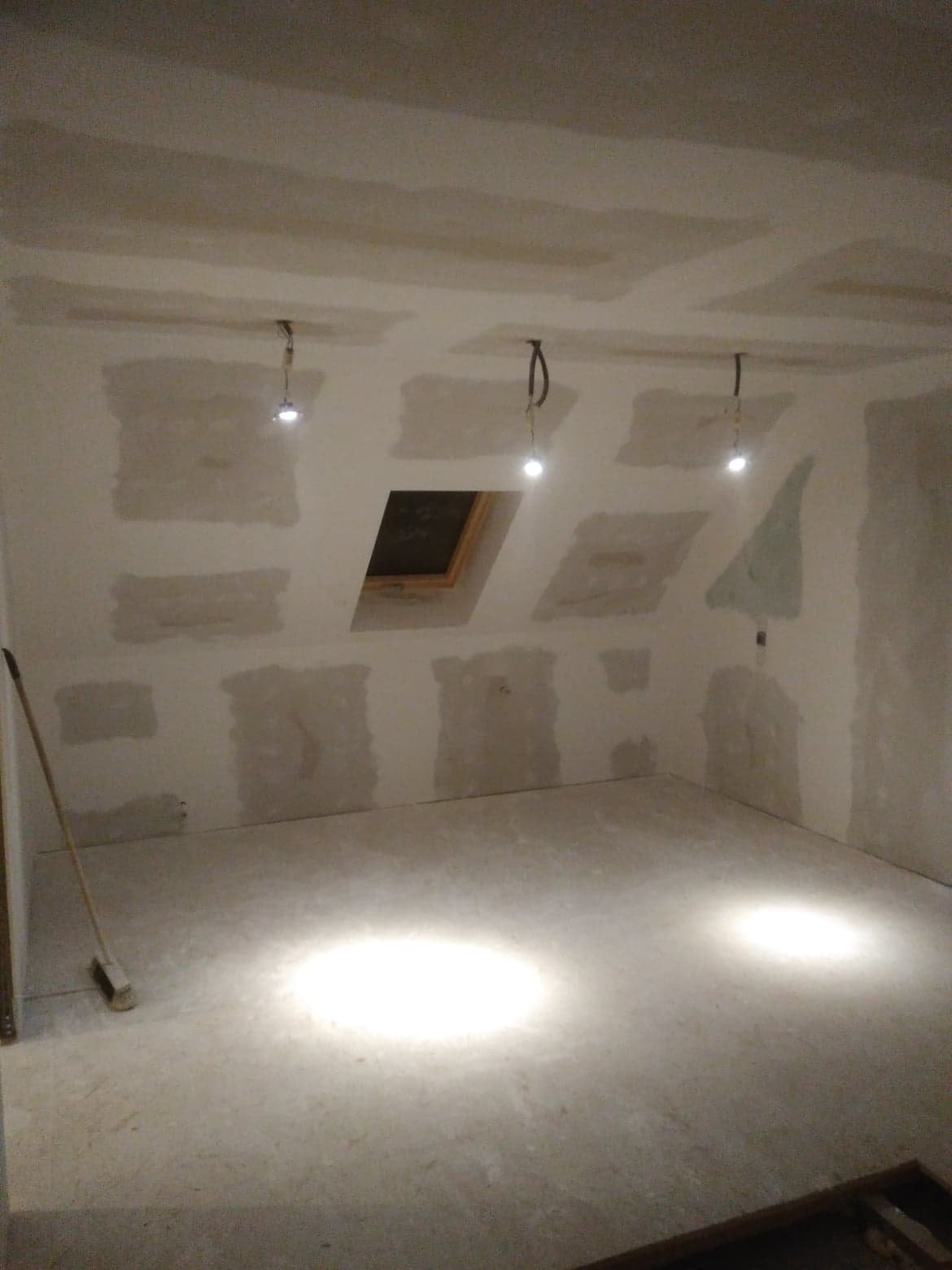 Pokój na poddaszu w trakcie wykańczania: ściany i sufit pokryte płytami gipsowo-kartonowymi, widoczne łączenia wymagające szpachlowania, okno dachowe i tymczasowe oświetlenie na kablach.