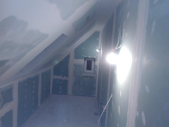 Poddasze w trakcie wykańczania, ściany i sufit pokryte zielonymi płytami gipsowo-kartonowymi, widoczne łączenia między płytami, małe okno z ciemną ramą, pomieszczenie oświetlone mocnym, punktowym...