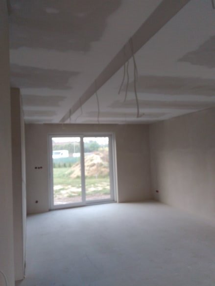 Wnętrze w trakcie prac wykończeniowych: widoczne płyty karton-gips na suficie z wystającymi przewodami elektrycznymi, szpachlowane ściany i wylewka na podłodze, okno balkonowe z widokiem na zewnątrz.