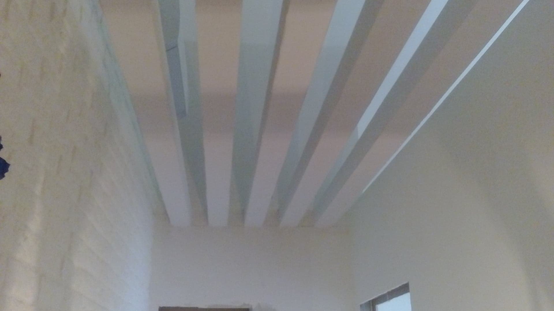 Białe, równoległe pasy na suficie, tworzące dekoracyjny element w pomieszczeniu z oświetleniem pośrednim i fragmentem ściany z fakturą cegły.