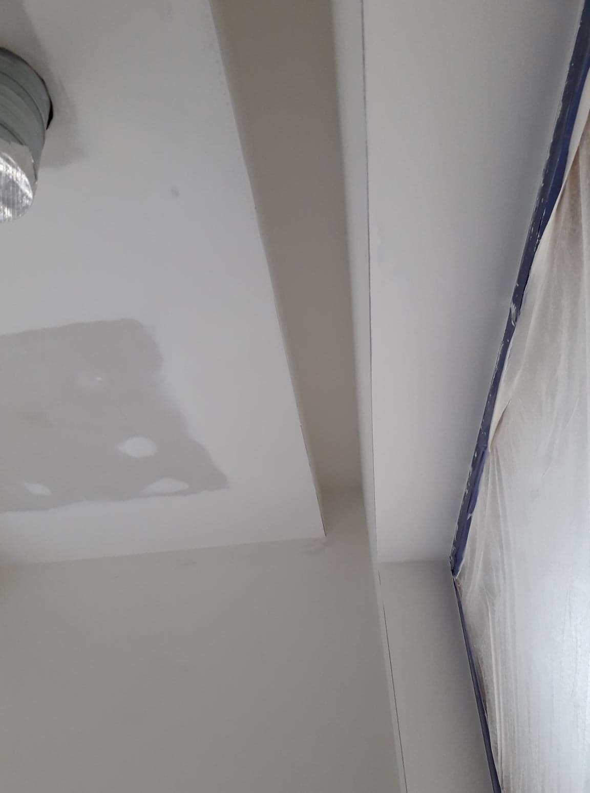 Fragment sufitu po szpachlowaniu, z widocznymi łatami i przygotowaniem do malowania, zabezpieczone folią okno w rogu.