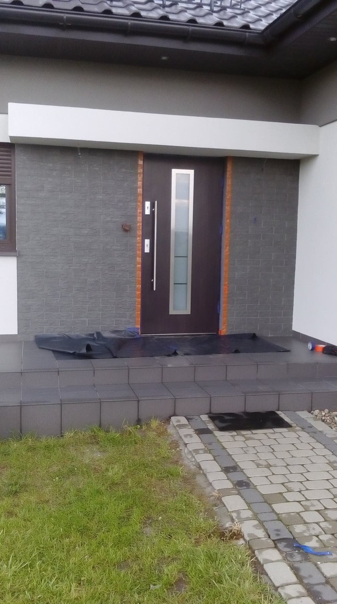 Elewacja zewnętrzna budynku jednorodzinnego z nowoczesnymi drzwiami wejściowymi, zabezpieczona folią malarską podczas prac wykończeniowych.