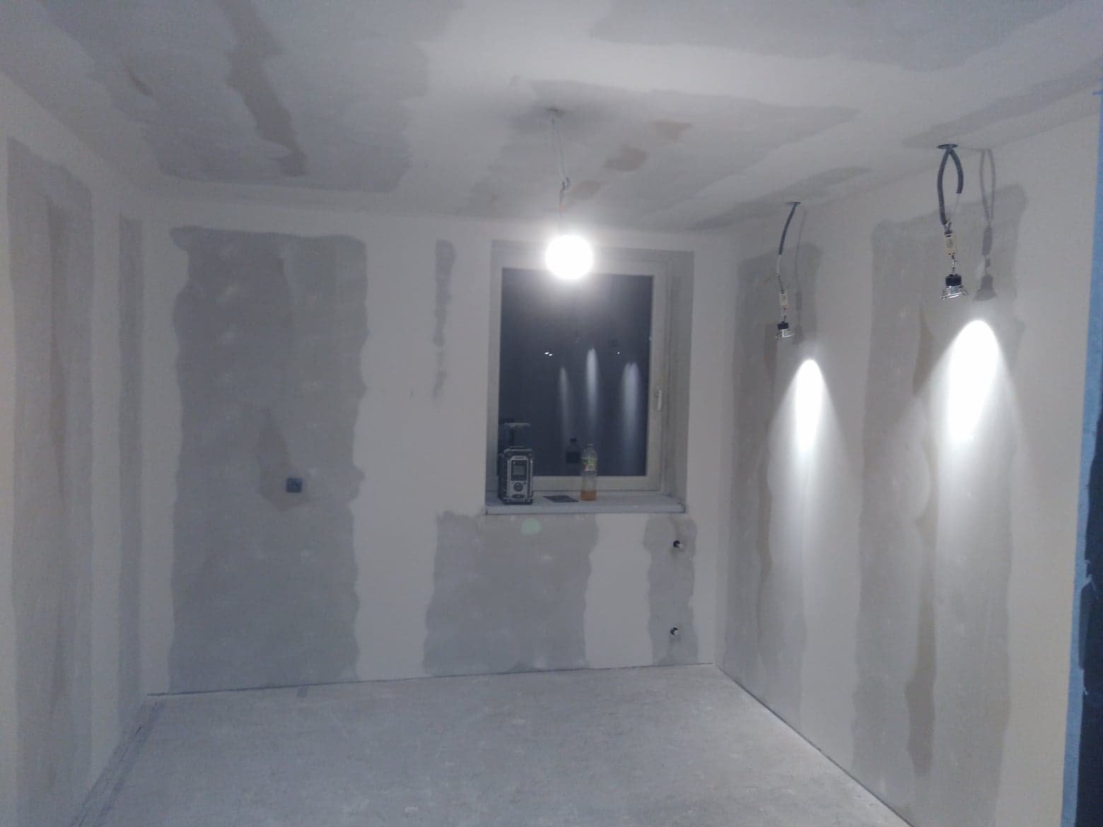 Pokój w trakcie remontu, ściany z częściowo nałożoną gładzią gipsową, widoczne przewody elektryczne, okno z butelką i dalmierzem laserowym.