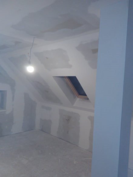 Pokój na poddaszu w trakcie wykańczania: ściany i sufit pokryte płytami gipsowo-kartonowymi, widoczne łączenia wymagające szpachlowania, okno dachowe w skosie dachu, surowa żarówka zwisająca...