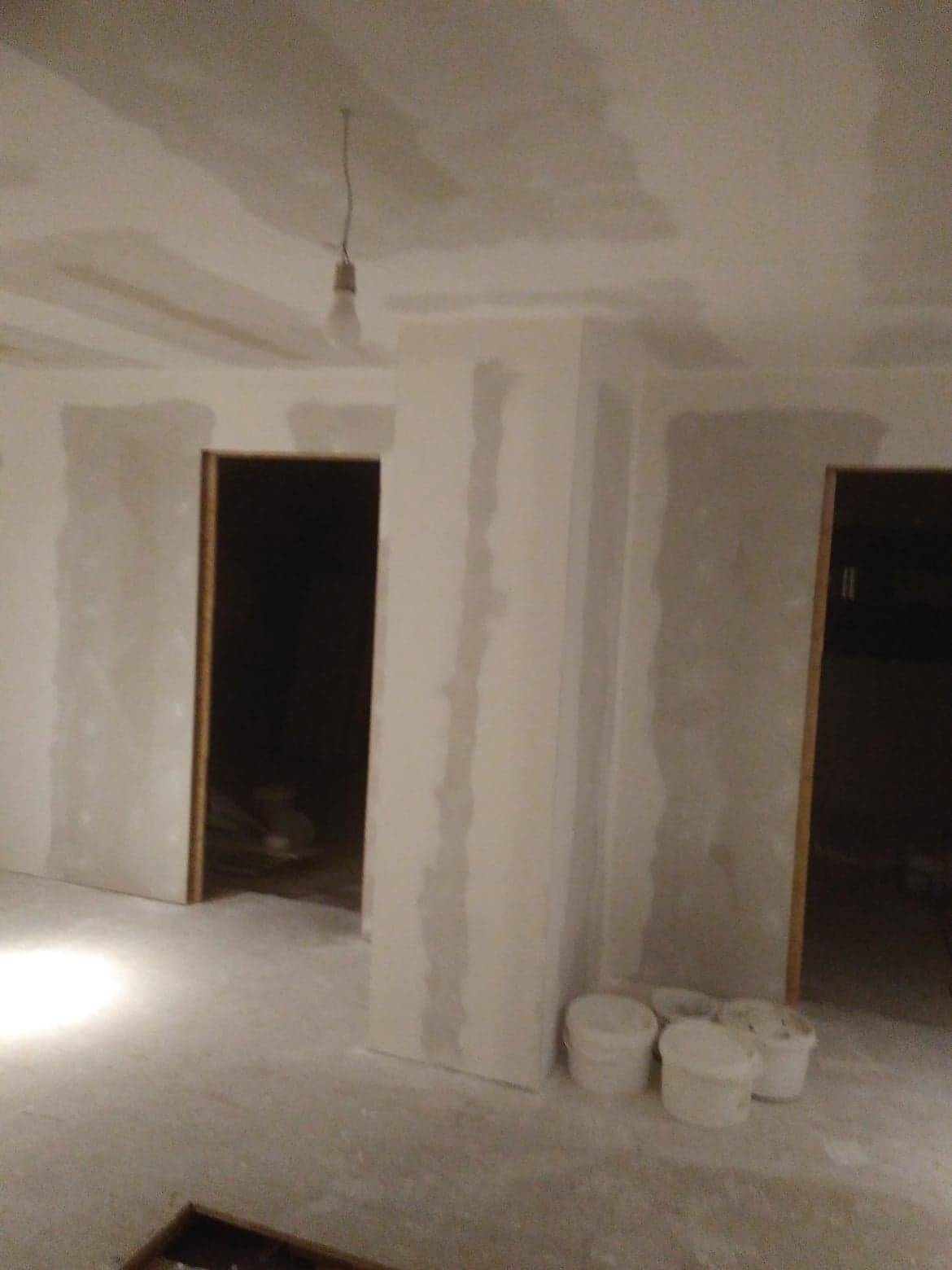 Wnętrze w trakcie prac wykończeniowych: zamontowane płyty gipsowo-kartonowe na ścianach i suficie, widoczne szpachlowane łączenia, surowa żarówka zwisająca z sufitu, oraz kilka plastikowych wiader...