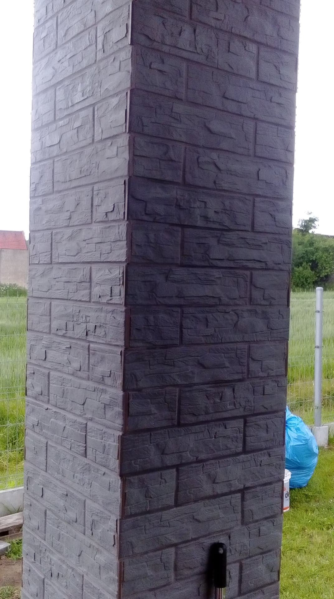 Wykończony narożnik budynku obłożony szarą imitacją kamienia, z widocznym fragmentem trawnika i niebieskim workiem w tle.