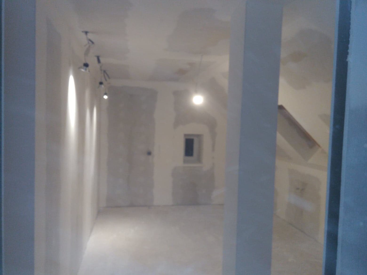 Wnętrze w trakcie prac wykończeniowych: ściany z widocznymi łączeniami płyt, sufit przygotowany do dalszych prac, oświetlenie punktowe na ścianie, małe okno w tle.