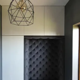 Wnęka z szarym, pikowanym obiciem w zabudowie meblowej, oświetlona lampą z geometrycznym kloszem.