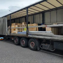 Ciężarówka z otwartą naczepą załadowana kartonami i meblami, widoczna z boku na parkingu.