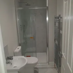 Nowoczesna łazienka z kabiną prysznicową wykończoną płytkami imitującymi marmur, białą umywalką z szafką, toaletą i chromowanym grzejnikiem drabinkowym.