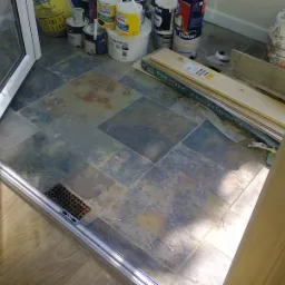 Podłoga z płytek w odcieniach szarości i brązu, z widoczną kratką wentylacyjną przy progu drzwi balkonowych, obok puszki farb i opakowania paneli podłogowych Krono Original.