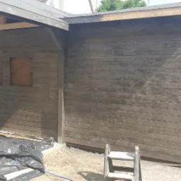 Malowanie natryskowe drewnianej konstrukcji w trakcie budowy; widoczny pracownik na drabinie, zabezpieczone okna, czarna folia ochronna na ziemi.