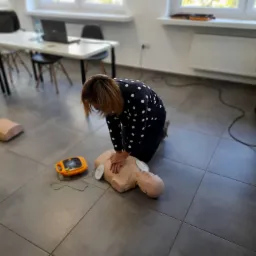 Kobieta w koszuli w kropki klęczy nad manekinem i wykonuje resuscytację krążeniowo-oddechową, obok leży defibrylator AED. W tle widoczne stoły, krzesła i okno.