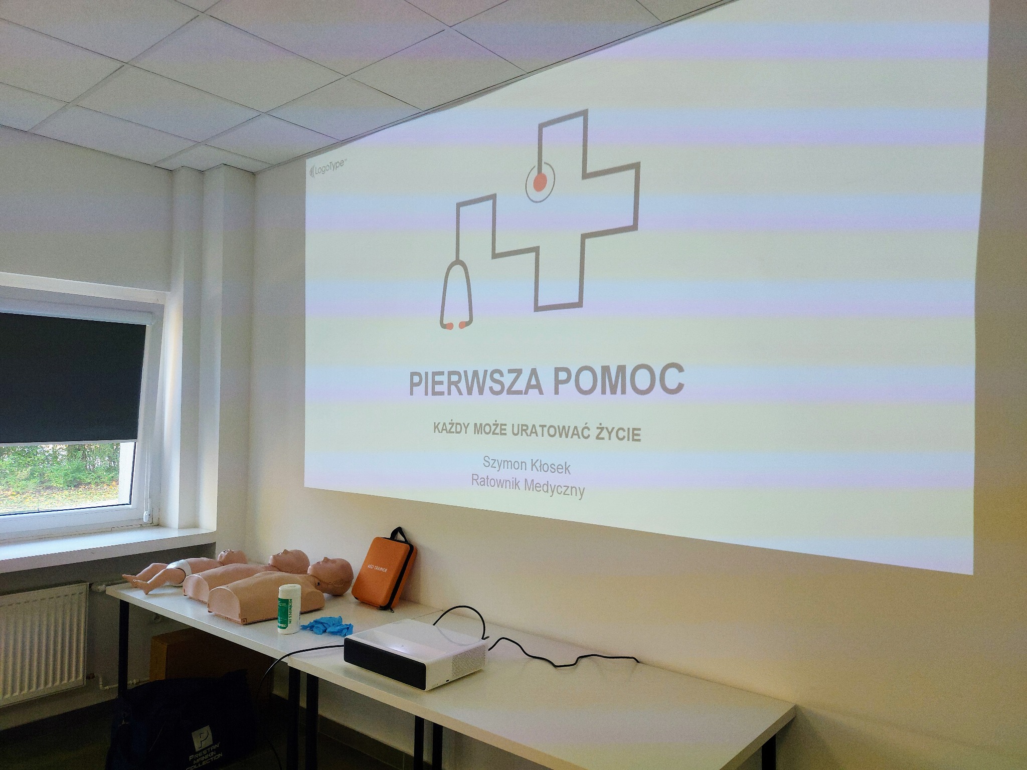 Sala szkoleniowa z manekinami do nauki pierwszej pomocy, projektorem wyświetlającym prezentację z logo i napisem 'PIERWSZA POMOC', butelką płynu dezynfekcyjnego i rękawiczkami.
