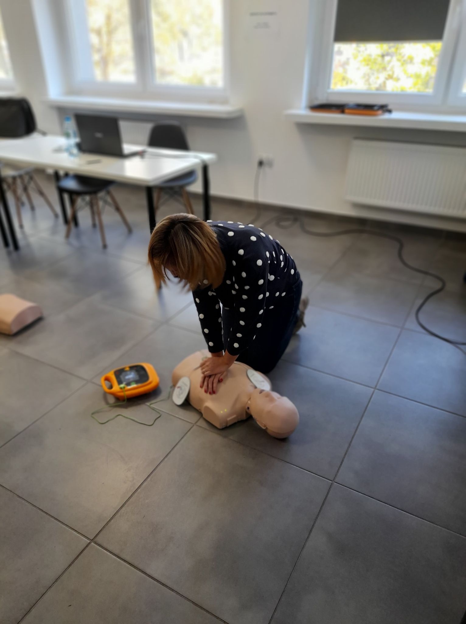 Kobieta w koszuli w kropki klęczy nad manekinem i wykonuje resuscytację krążeniowo-oddechową, obok leży defibrylator AED. W tle widoczne stoły, krzesła i okno.
