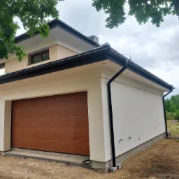 Nowoczesny budynek z brązową bramą garażową i jasną elewacją, widoczne rynny w kolorze czarnym, fragment drzewa w górnym rogu kadru, teren wokół budynku niezagospodarowany.