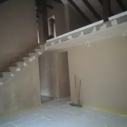 Surowe wnętrze poddasza z drewnianą konstrukcją dachu, ściany pokryte świeżym tynkiem, podłoga zabezpieczona styropianem, widoczne prowizoryczne oświetlenie i narzędzia