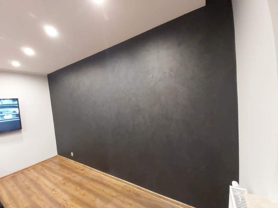 Ściana w pokoju pomalowana na ciemny grafit z widoczną fakturą, kontrastująca z jasną podłogą z paneli drewnopodobnych i białymi ścianami bocznymi, oświetlona punktowymi lampami sufitowymi.
