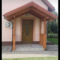 Wejście do domu z drewnianym zadaszeniem i balustradą, widoczne liczniki na elewacji, trawnik przed domem.